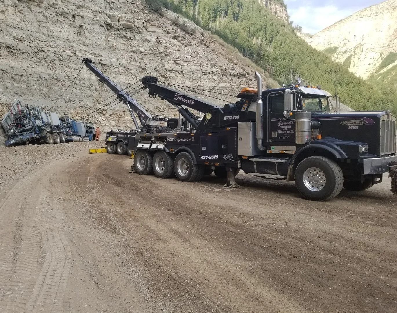 A large black heavy-duty tow truck parked on a dirt road, preparing to recover a wrecked vehicle near a rocky hillside.