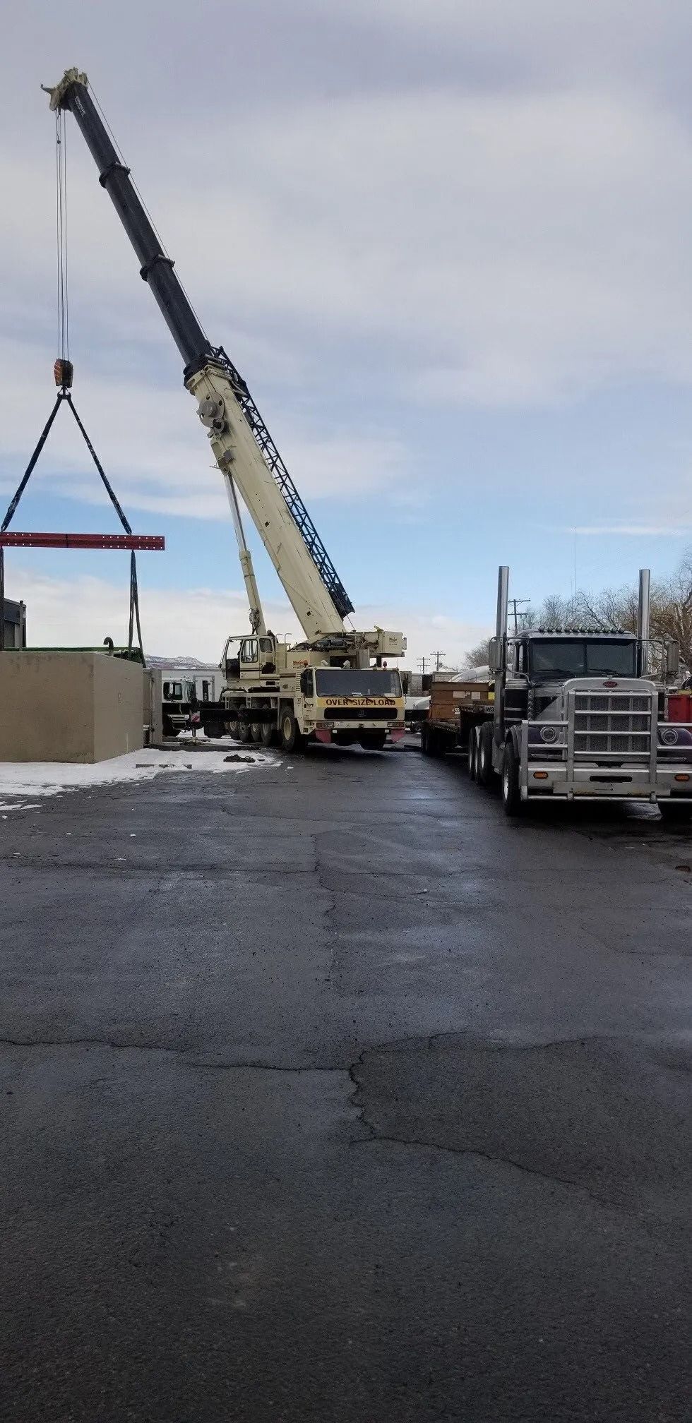 A large crane extends its boom over a beige building in a paved, snowy lot next to a parked white semi-truck.