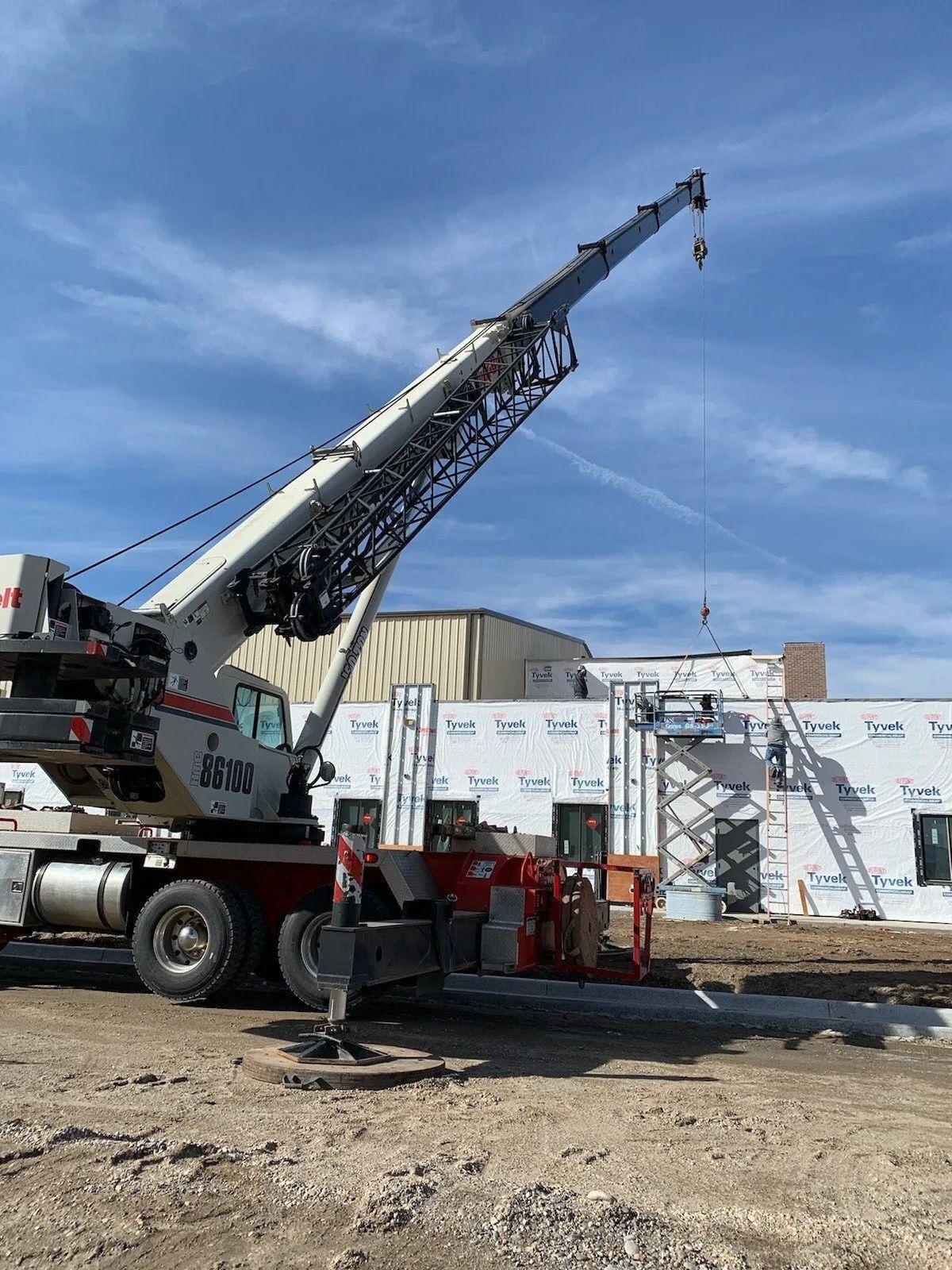 A crane sits at a construction site in front of a building wrapped in white protective sheeting under a clear blue sky.