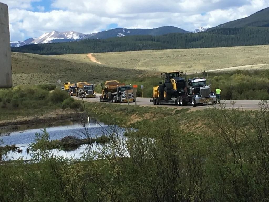 A line of semi-trucks hauling heavy construction machinery parked on a roadside near a pond with mountains in the background.