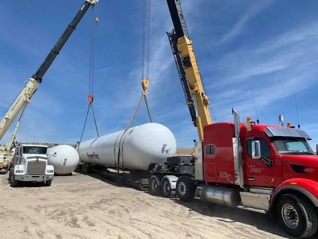 Crane lifting a large white tank onto a red semi truck in a sandy construction yard