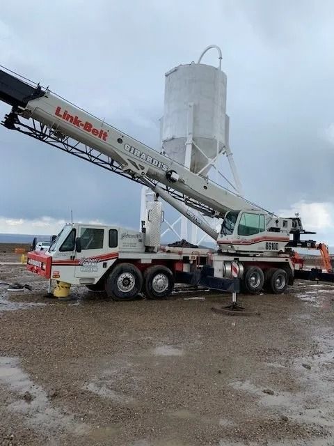 A white Link-Belt mobile crane parked on a gravel construction site with a tall, white industrial silo in the background.