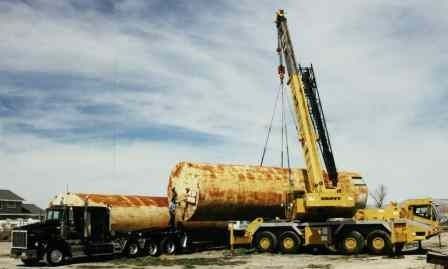 A yellow mobile crane lifts a large, rusty cylindrical tank near a black semi-truck outdoors under a cloudy sky.