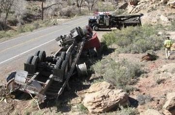 A semi-truck overturned on its side off a rural road, with a recovery tow truck parked nearby and a person observing.