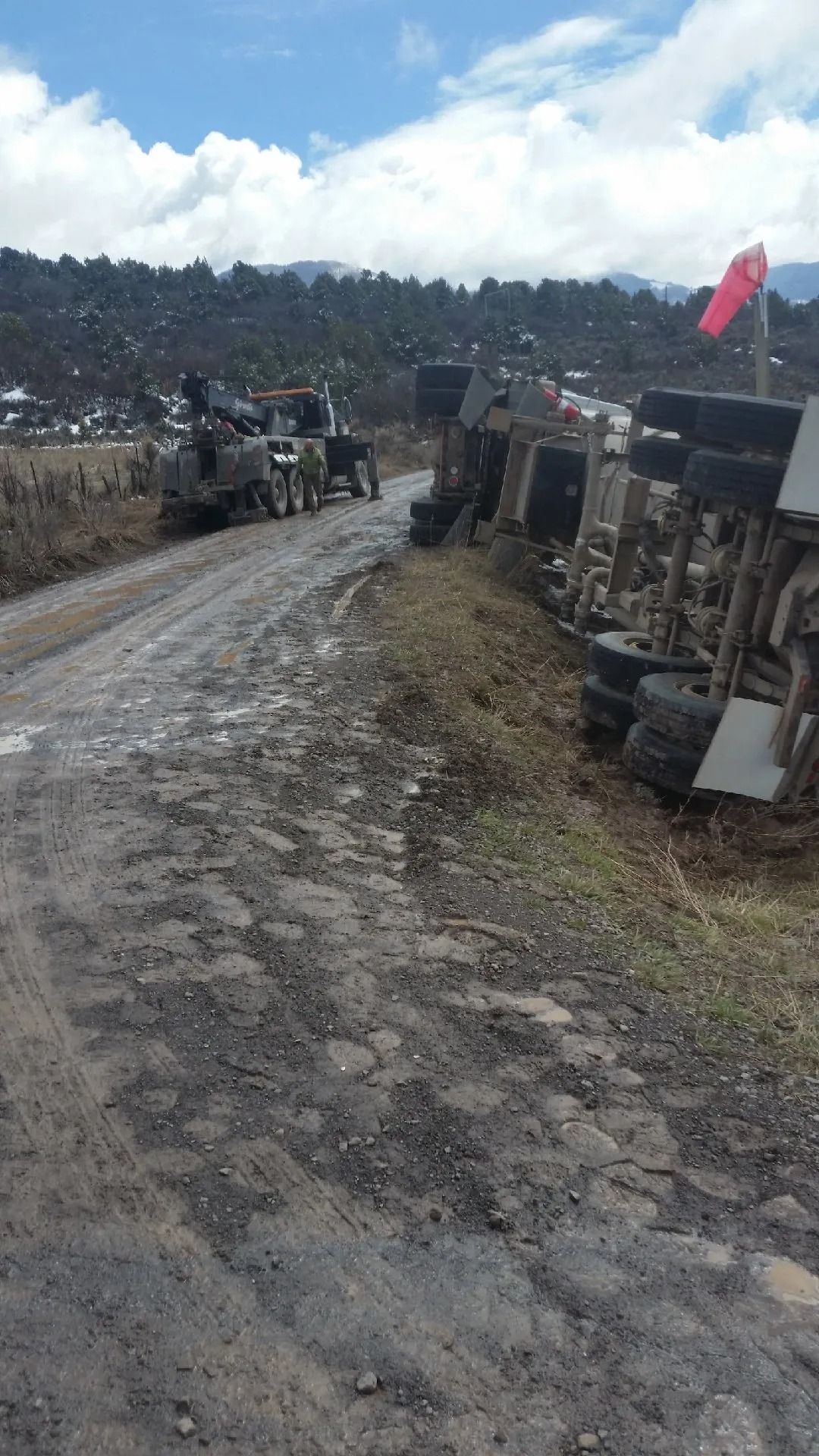 A large truck lies overturned on its side along a muddy, rural road, with a recovery vehicle parked nearby.