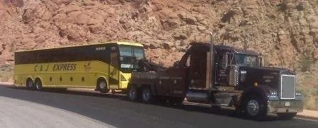 A black tow truck towing a yellow C&J Express tour bus along a road with a rocky cliff in the background.