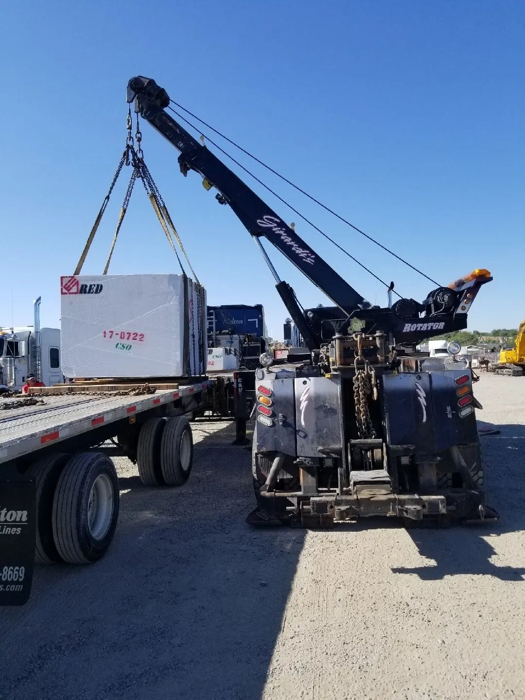 Crane lifting a large white container onto a flatbed truck in an industrial yard under a clear blue sky