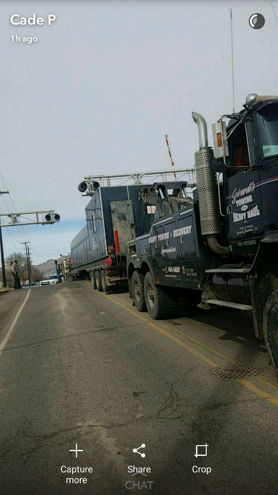 A dark-colored heavy-duty tow truck pulling a large trailer on a paved road under a cloudy sky.