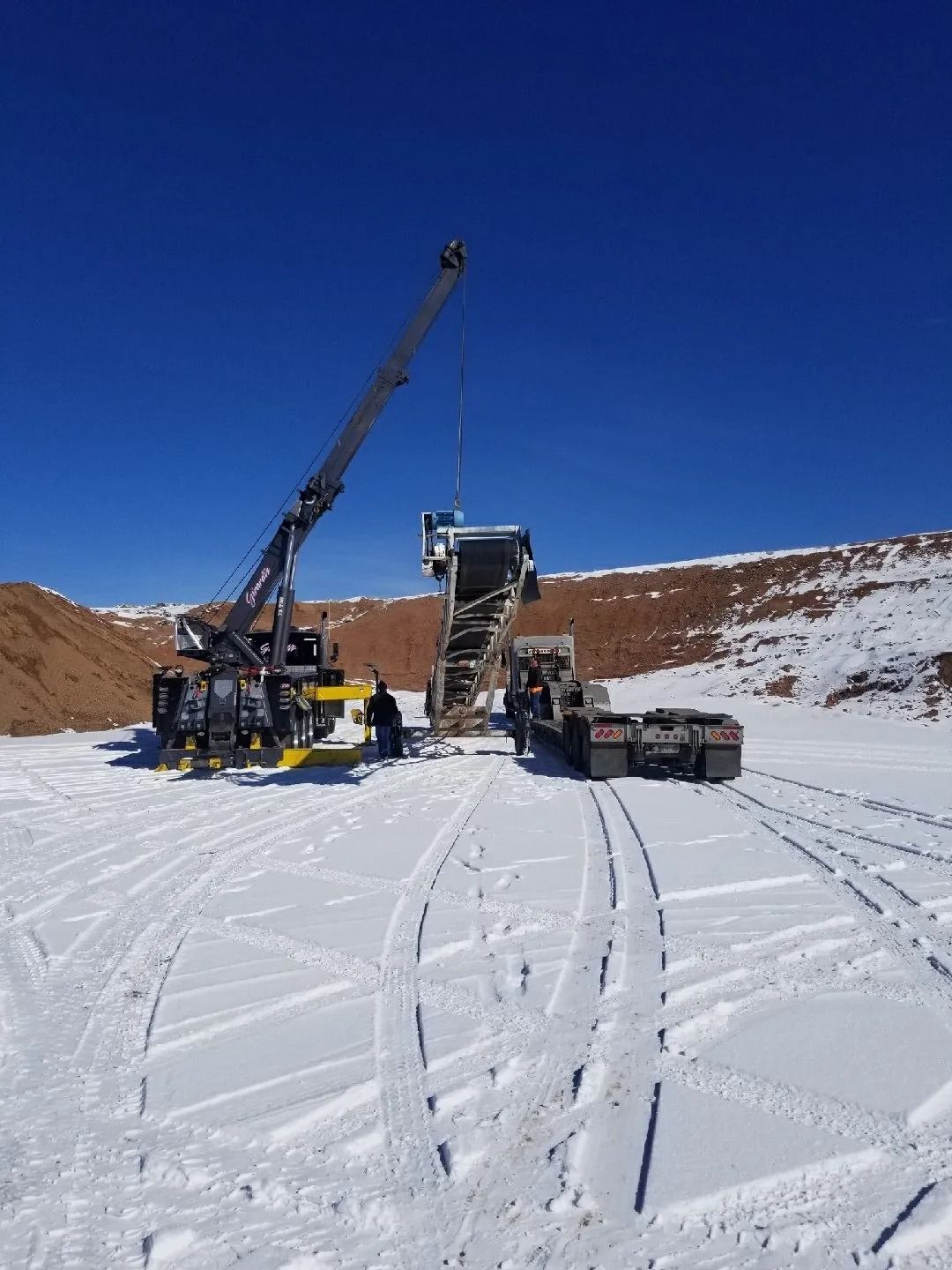 A crane lifts a large piece of industrial machinery onto a flatbed trailer in a snow-covered quarry under a clear blue sky.