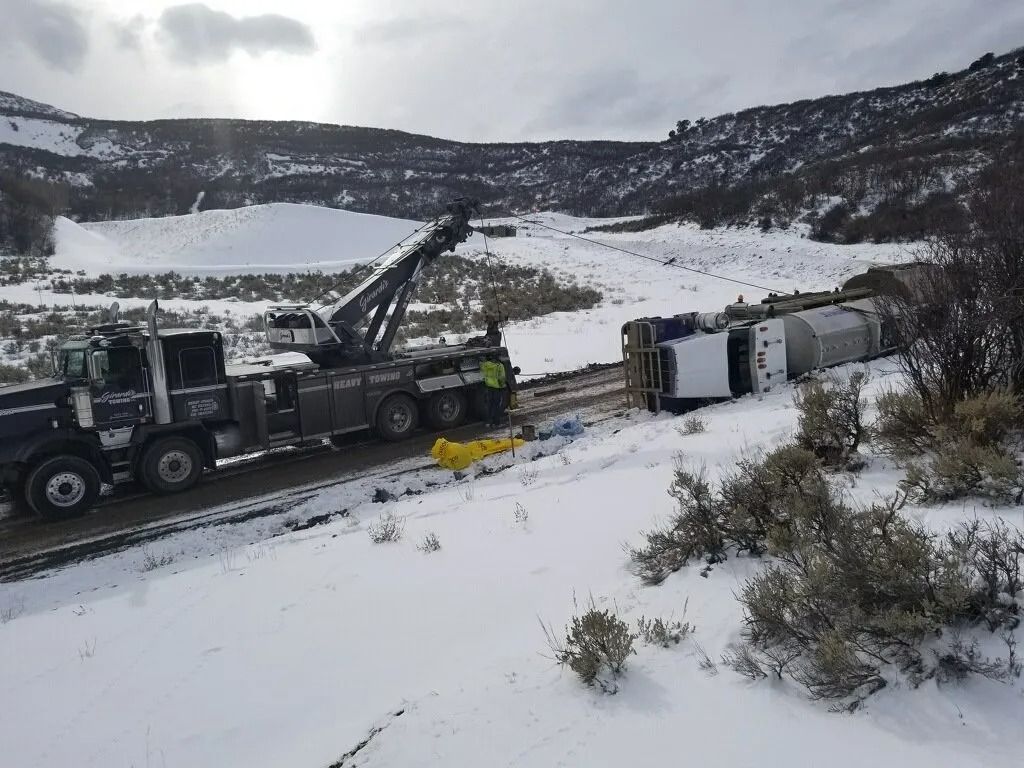 A heavy-duty tow truck is recovering a white tanker truck that has overturned on a snow-covered, rural road.