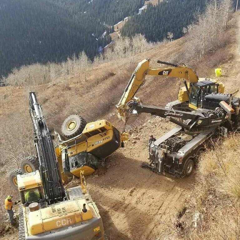 A yellow excavator is tipped over on a dirt hillside, being stabilized by a large heavy-duty recovery tow truck.