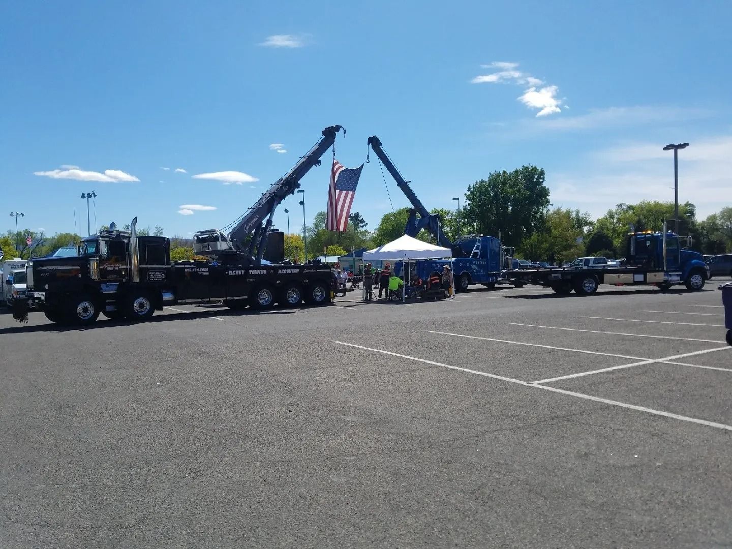 Two large tow trucks in a parking lot hold an American flag between their extended booms above a white event tent.