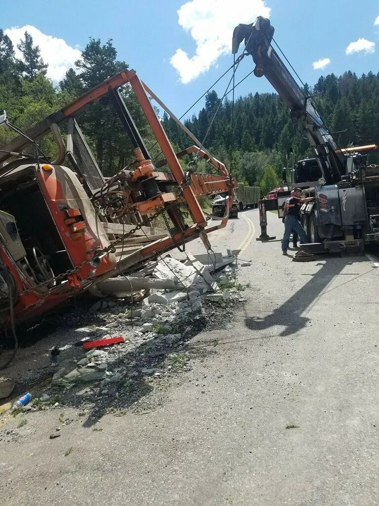 A tow truck uses a boom to lift an overturned orange piece of heavy machinery off a roadside.