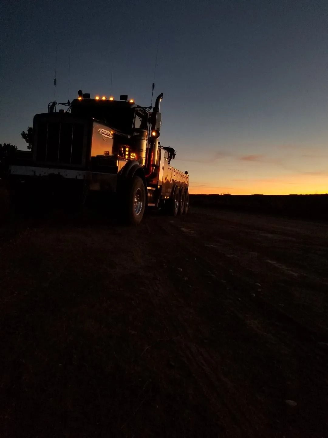 A large tow truck silhouette stands on a dirt road during a sunset with an orange horizon.