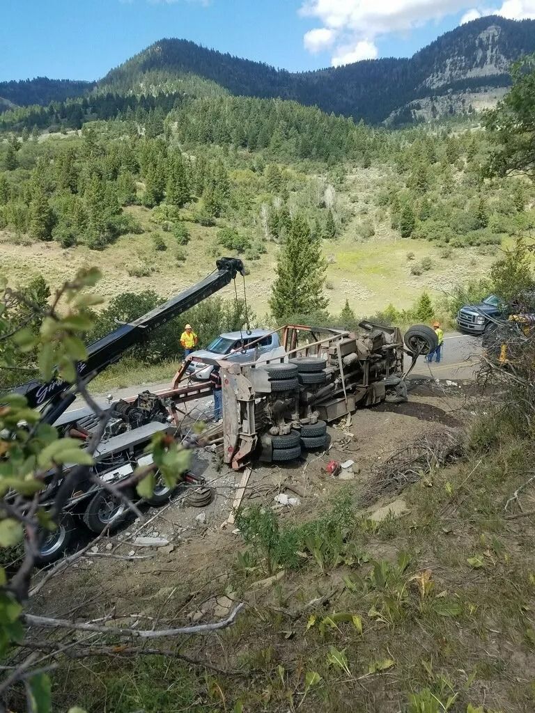 A tow truck works to recover a large vehicle that has overturned on a dirt road in a mountainous, forested area.