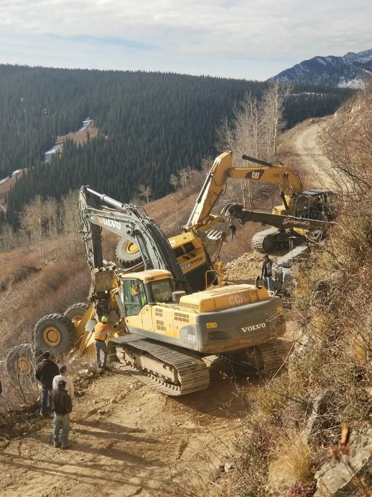 Two yellow excavators are on a dirt mountain road; one is lifting the other’s tracks, with workers standing nearby.