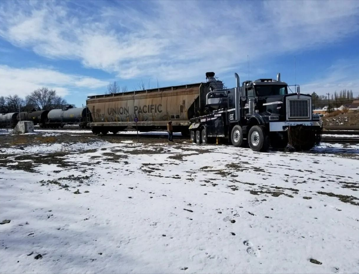 A dark semi-truck is parked on a snowy lot next to a large Union Pacific freight train car under a blue, cloudy sky.