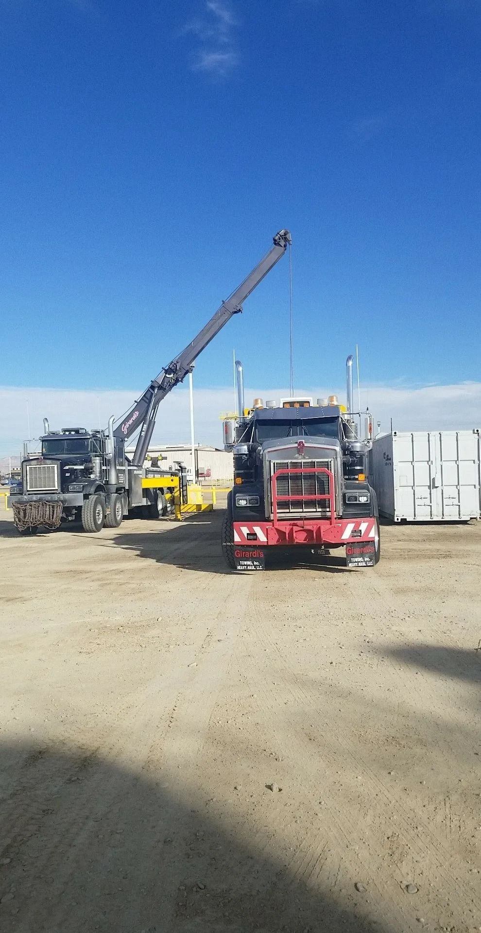 A grey tow truck with an extended crane arm positioned next to a parked semi-truck on a gravel lot under a blue sky.