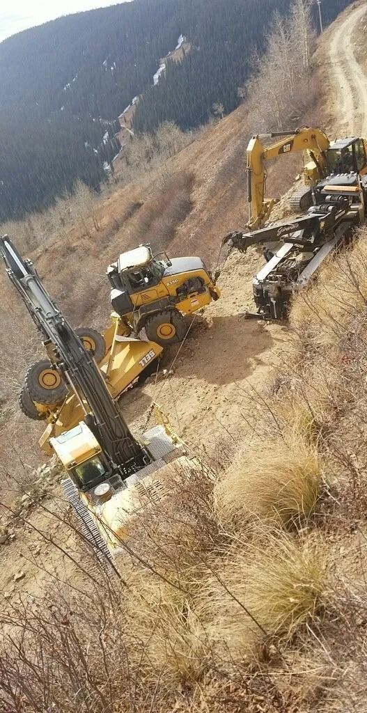 Two yellow construction vehicles, including one with a tall drilling rig, on a steep, dirt hillside path.