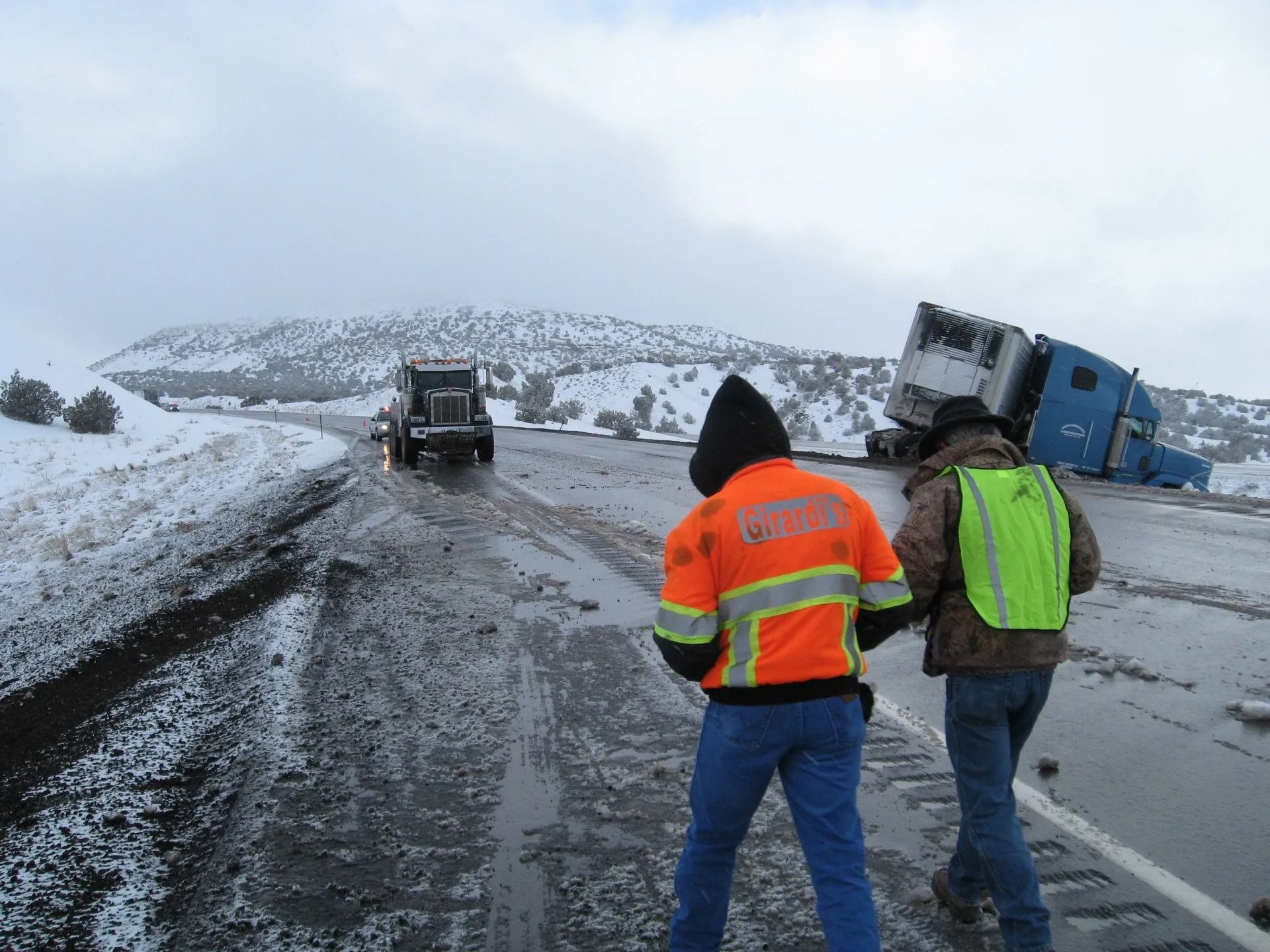 Two people in high-visibility safety gear walk toward a semi-truck that has overturned on a snowy, wet road.