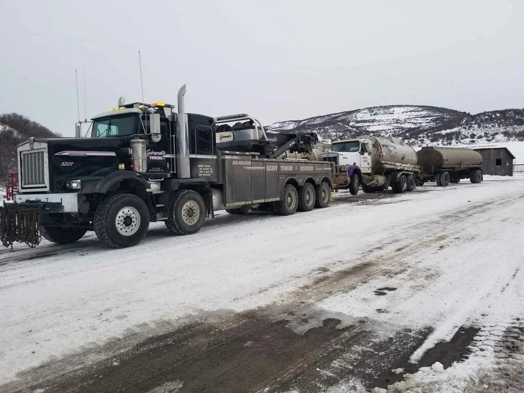 A black heavy-duty tow truck is pulling a tanker trailer along a snowy, rural road with mountains in the background.