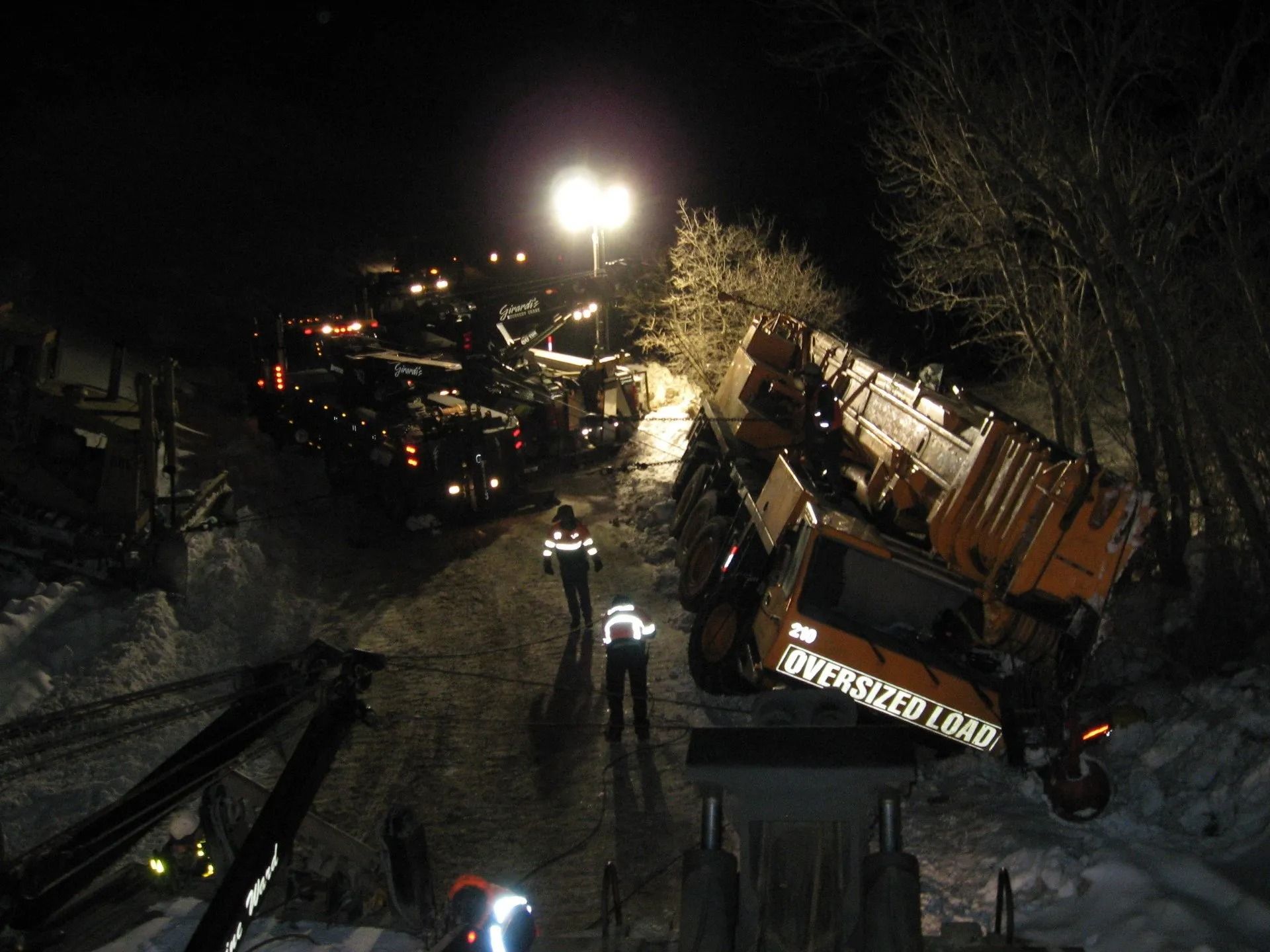 An orange oversized crane lies tipped on its side in a snowy, dark construction area as workers gather nearby.