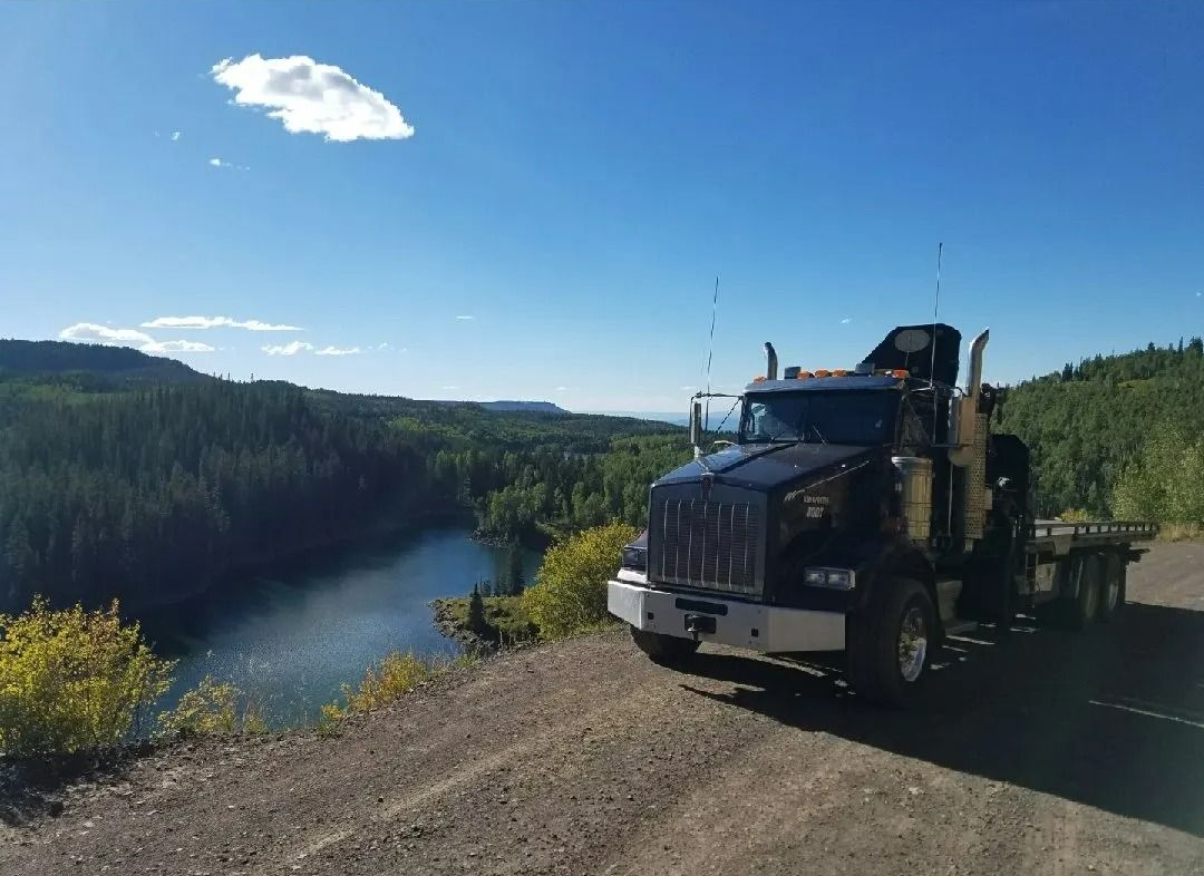 A black semi-truck is parked on a dirt cliff overlooking a lake surrounded by forest under a clear blue sky.