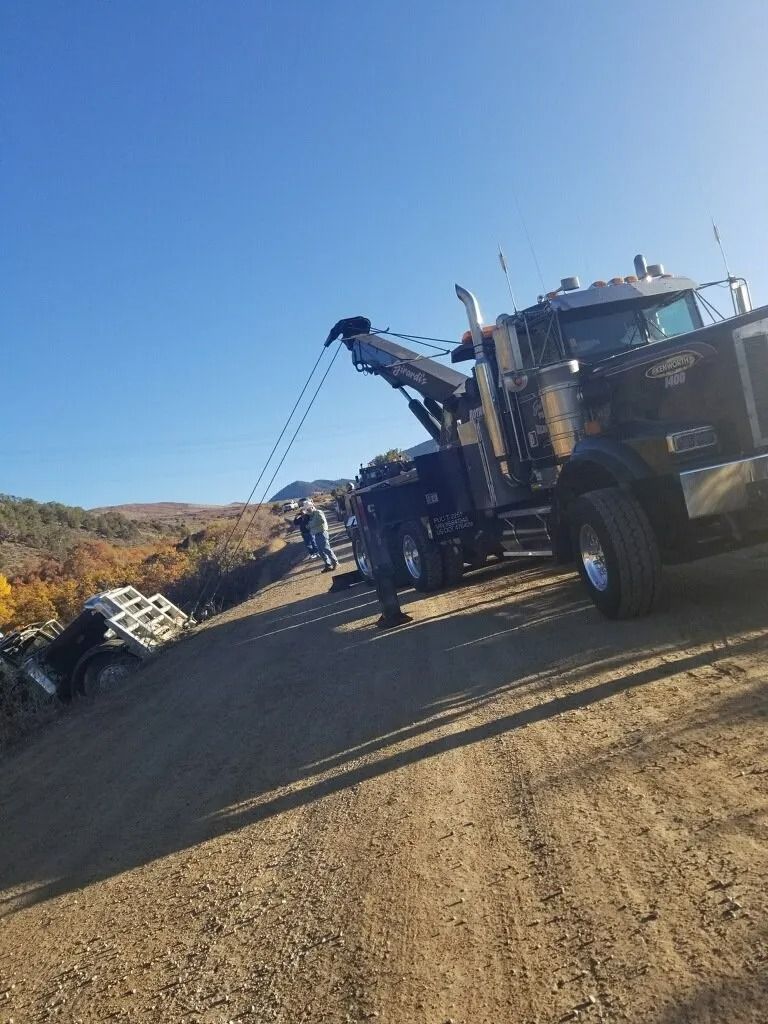 A large dark tow truck positioned on a dirt road, extending its boom to recover a white vehicle down an embankment.