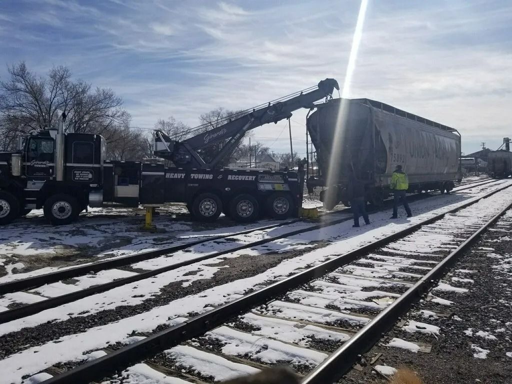 A heavy-duty tow truck lifts a derailed train car from snowy railroad tracks as workers stand nearby.