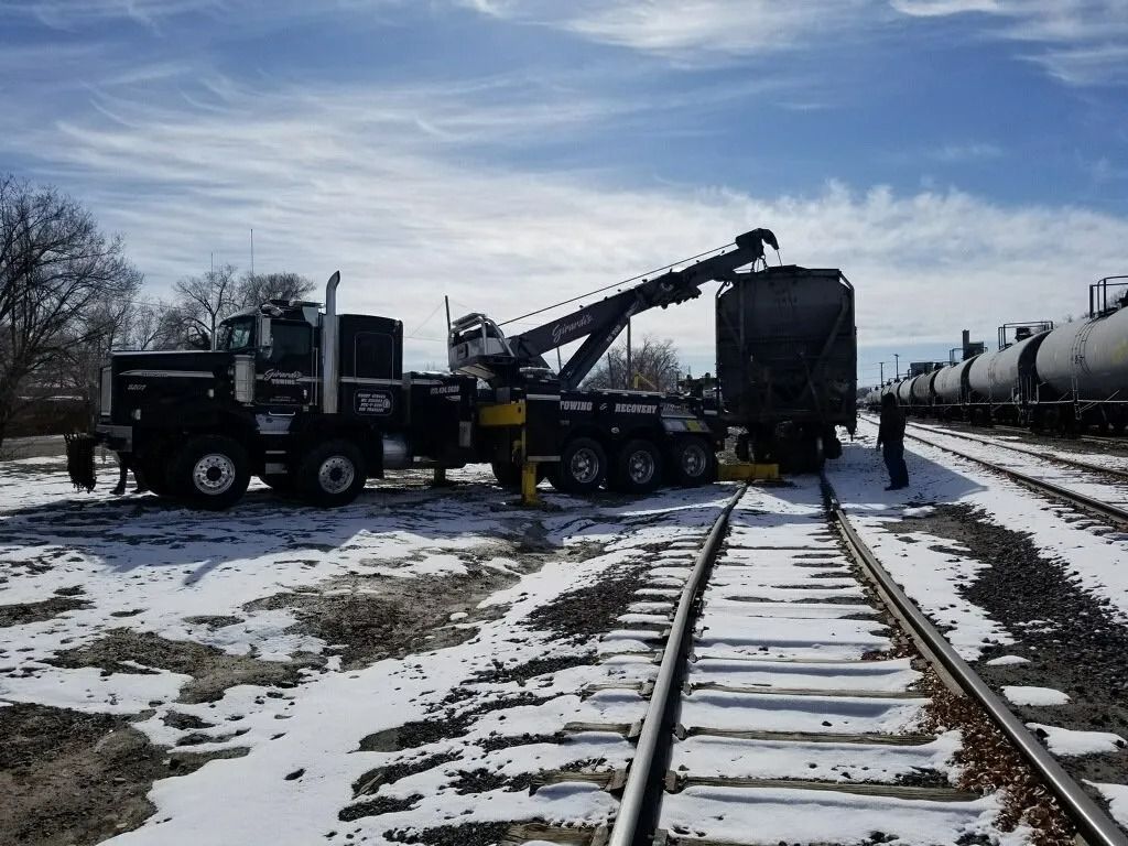 A black heavy-duty tow truck is using its crane arm to lift a train car on a snowy, industrial railroad track.