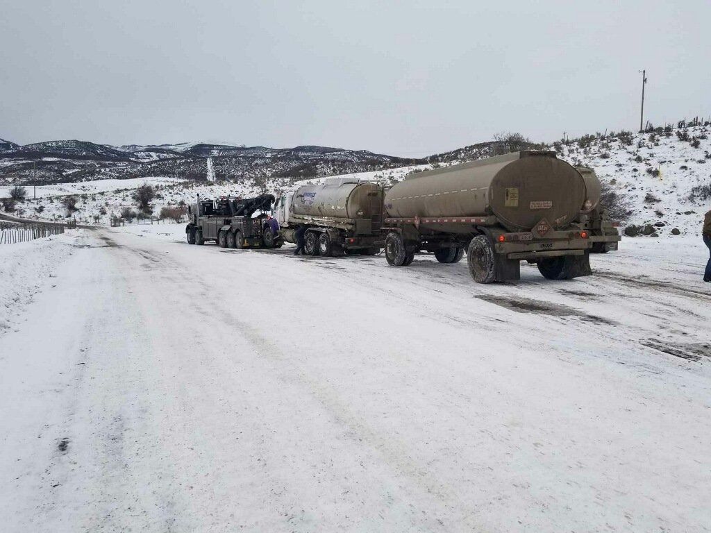 A tow truck pulls two tanker trailers along a snow-covered, rural road under a gray, overcast sky.
