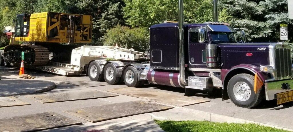 A purple semi-truck pulling a flatbed trailer loaded with a large yellow construction vehicle on a paved road.