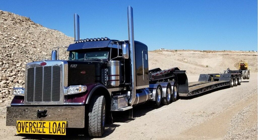 A dark purple Peterbilt semi-truck with an empty lowboy trailer parked in a gravel quarry under a clear blue sky.