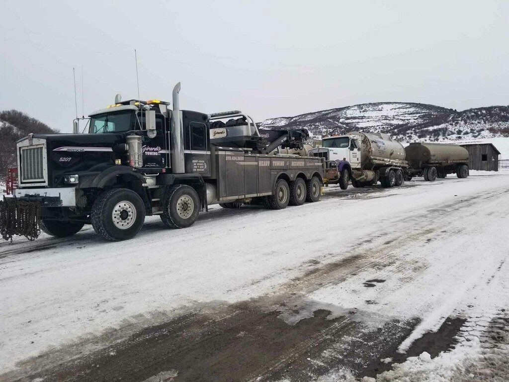 A black heavy-duty tow truck is hooked to a tanker truck on a snow-covered road in a mountainous, wintry landscape.
