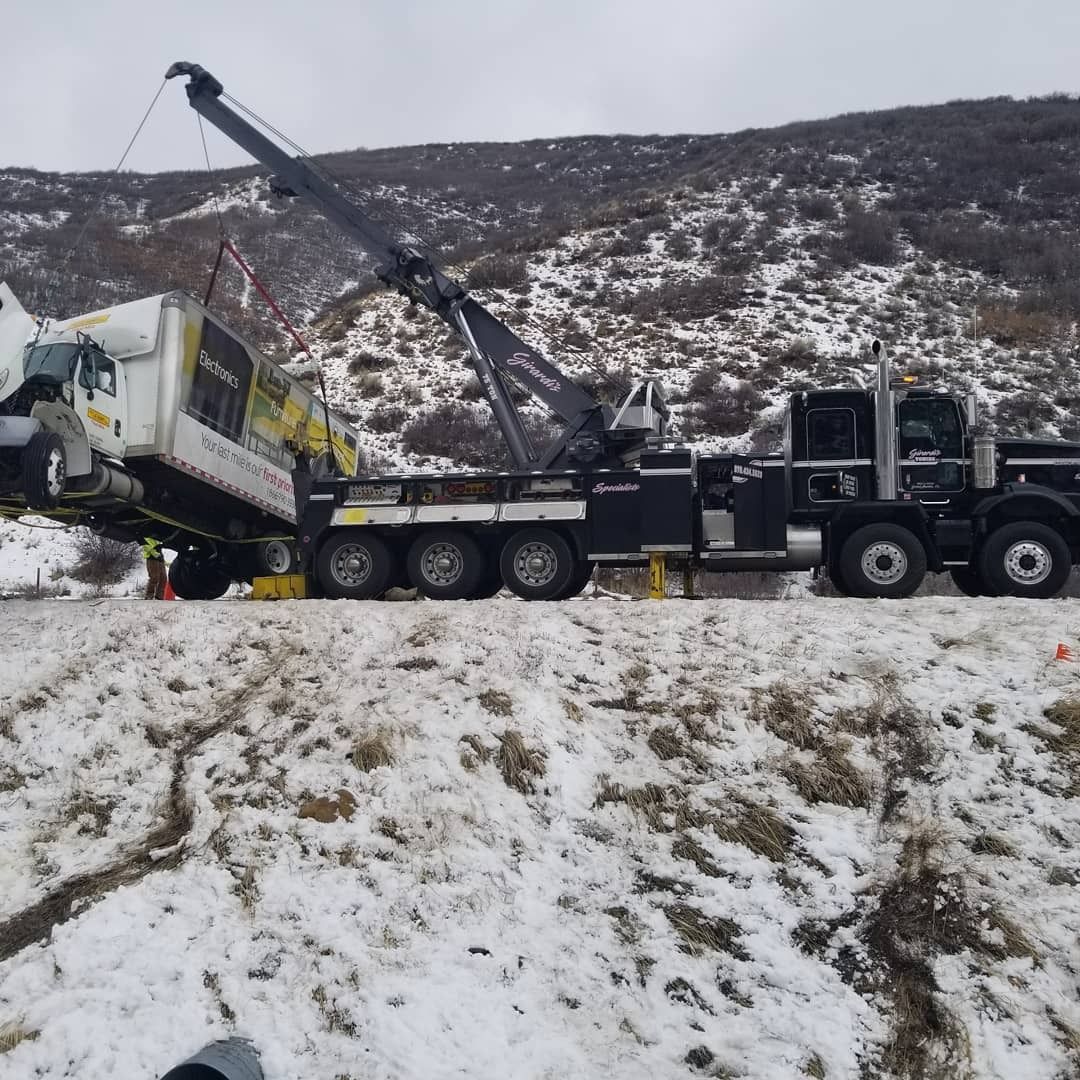 A black heavy-duty tow truck lifting a white delivery truck from a snowy, hillside roadside.