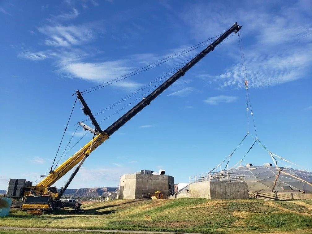 A large yellow mobile crane lifting a geodesic dome frame over concrete structures under a blue sky.