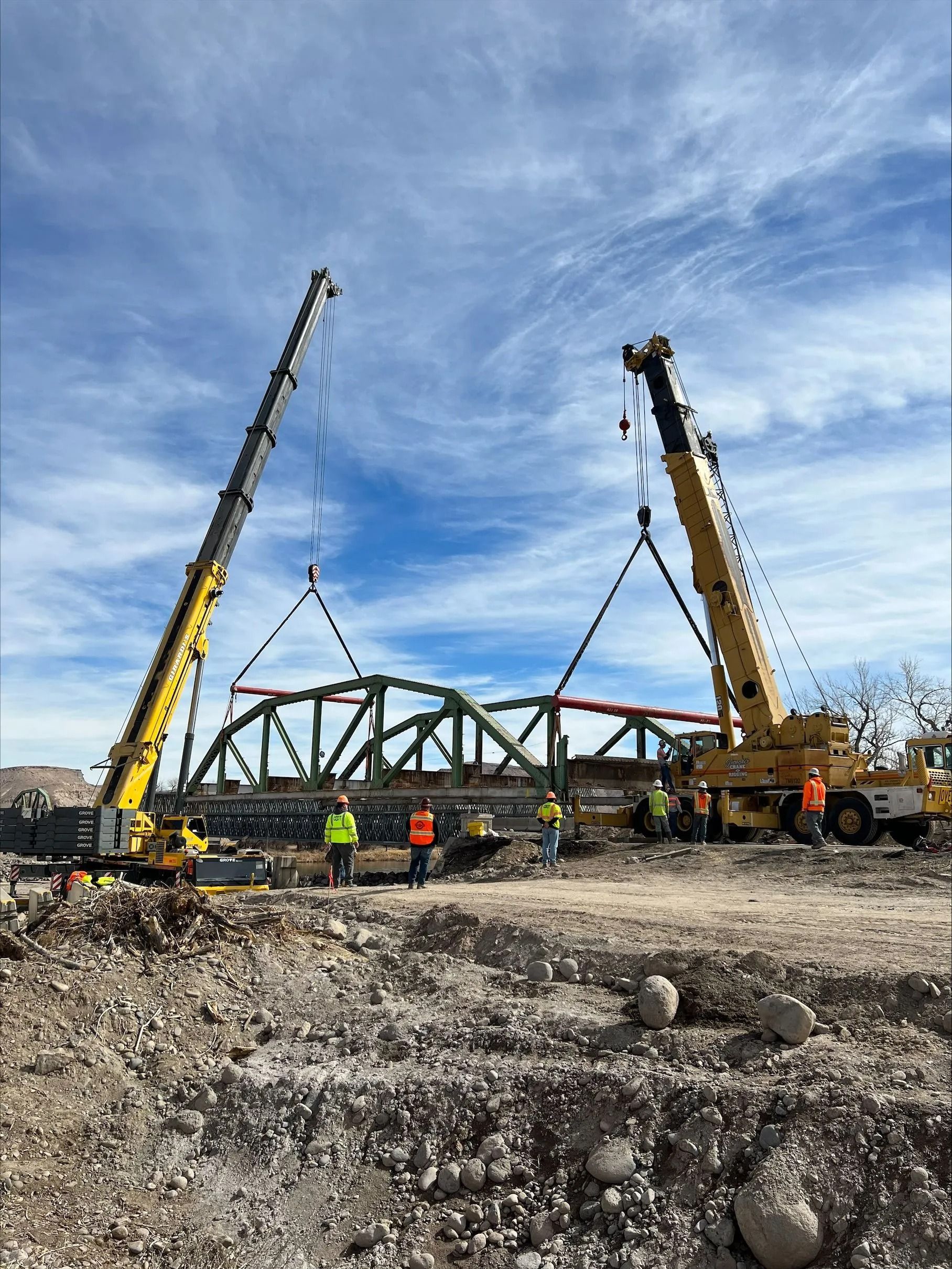Two yellow cranes lift a large green metal bridge truss over a dirt construction site under a blue, cloudy sky.