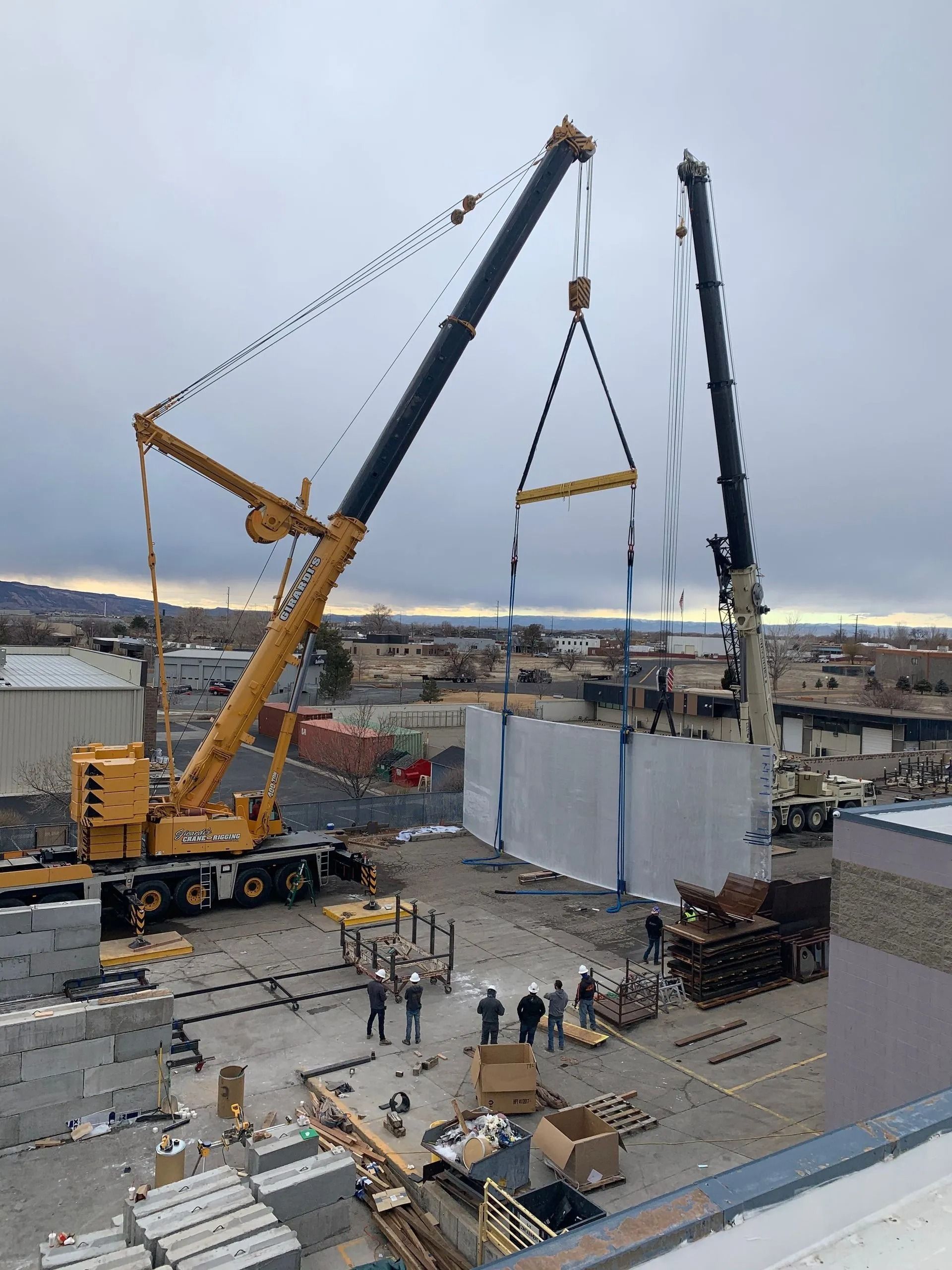 Two large yellow and white cranes collaborate to lift a concrete wall panel at a construction site with crew members nearby.