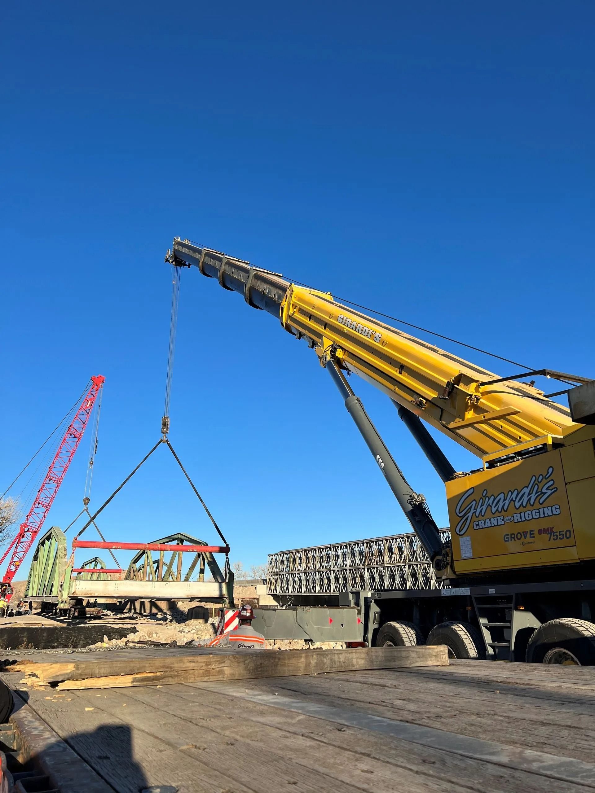 A yellow mobile crane lifts structural steel at a construction site against a clear blue sky.