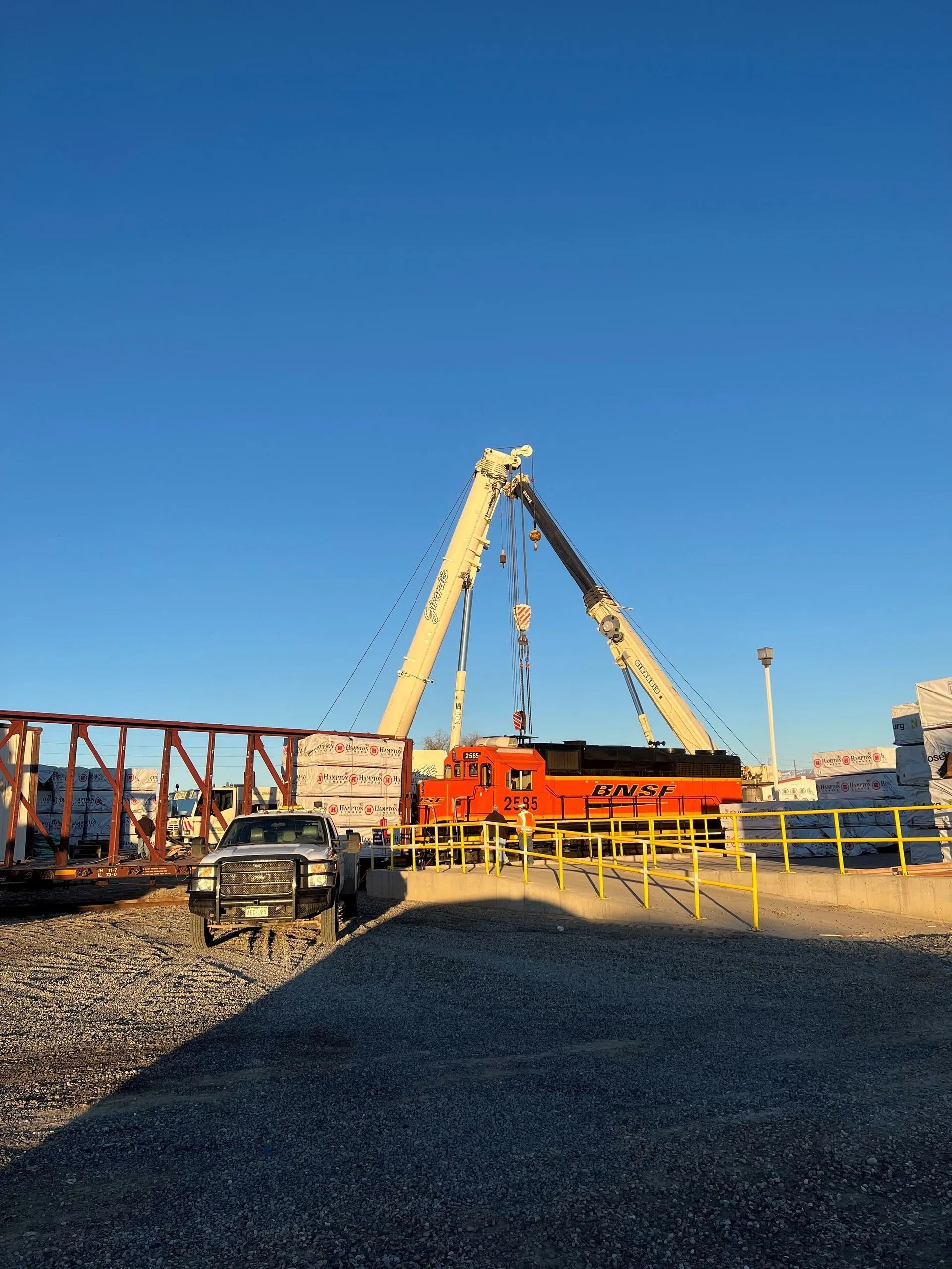 A large crane lifts a bright orange BNSF locomotive near a railroad bridge under a clear blue sky.