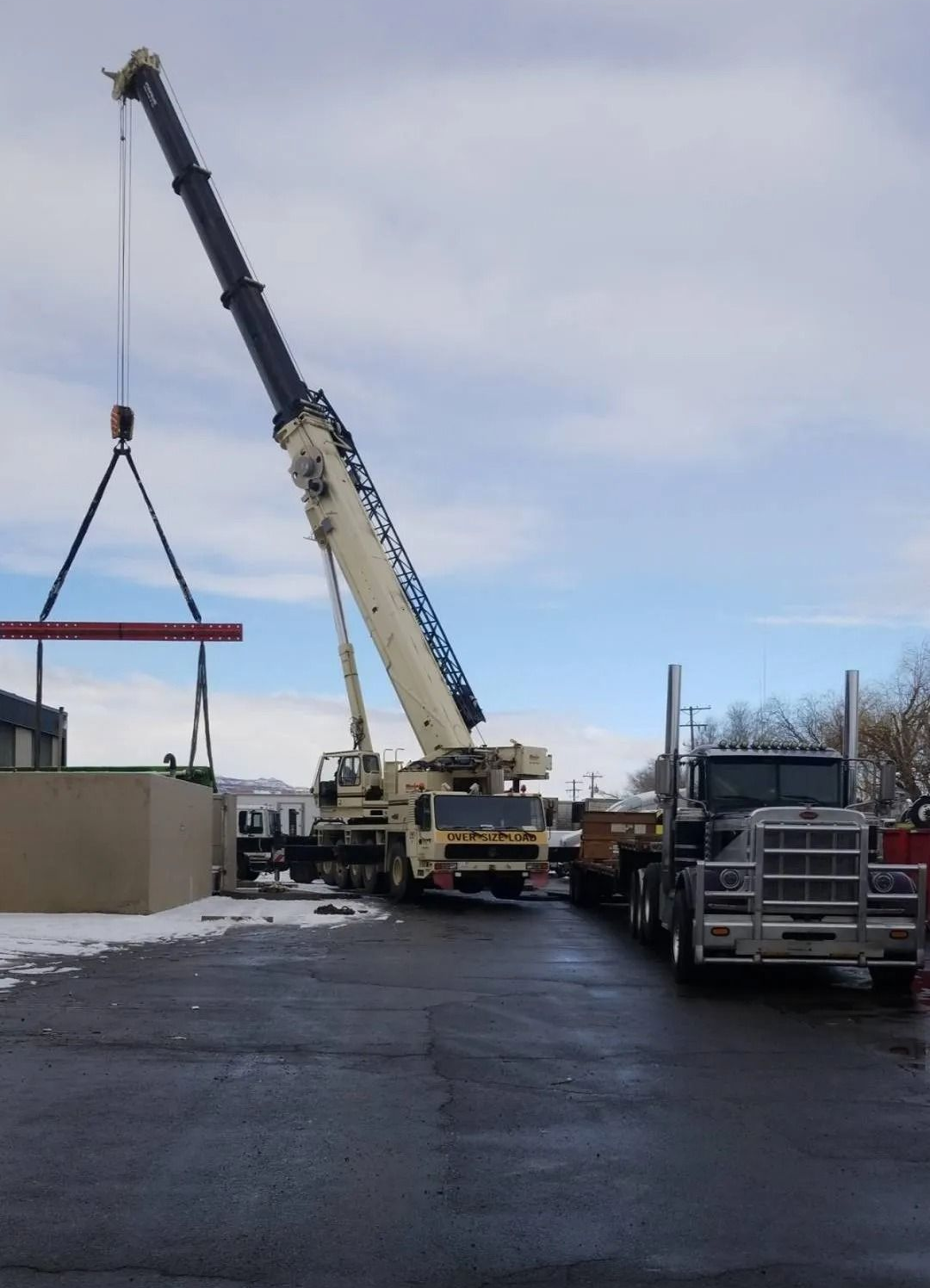 Crane lifting a long red beam at a construction site beside parked trucks.
