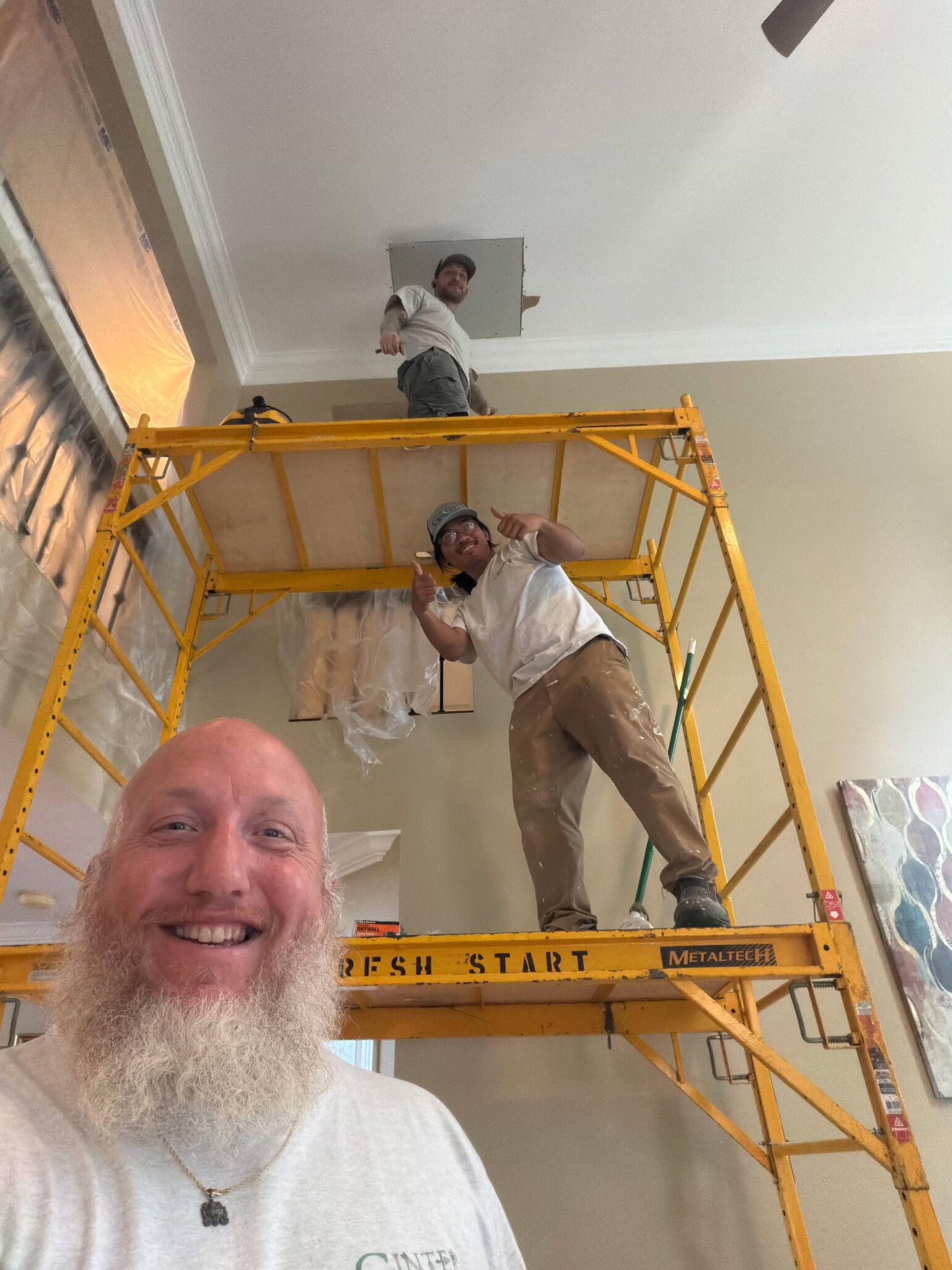 Three men on a yellow scaffold indoors, smiling during ceiling work in a bright room.