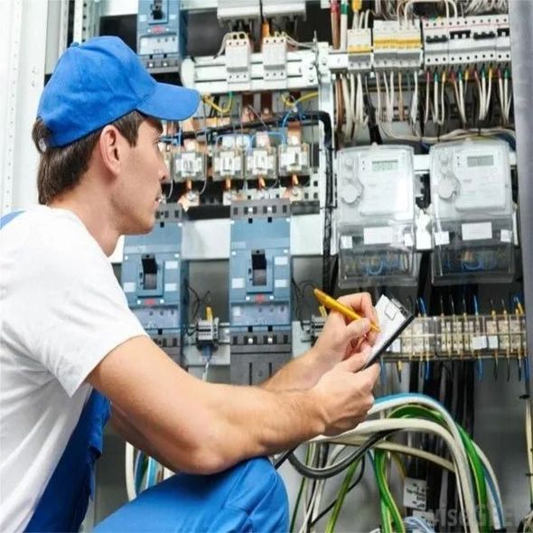 Electrician inspecting an open electrical panel while holding a yellow pencil, wearing a blue cap.