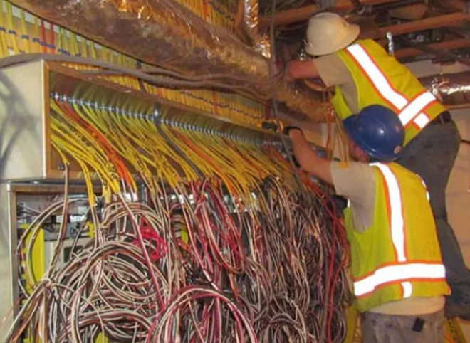 Two workers in hard hats inspect a dense bundle of cables in an industrial wiring room.