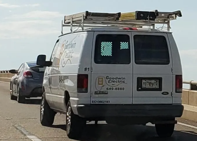 White utility van with ladder rack driving on a highway, with a car following behind