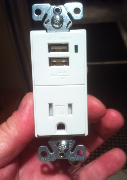 White wall outlet with two USB ports and a standard plug receptacle, held by hand.