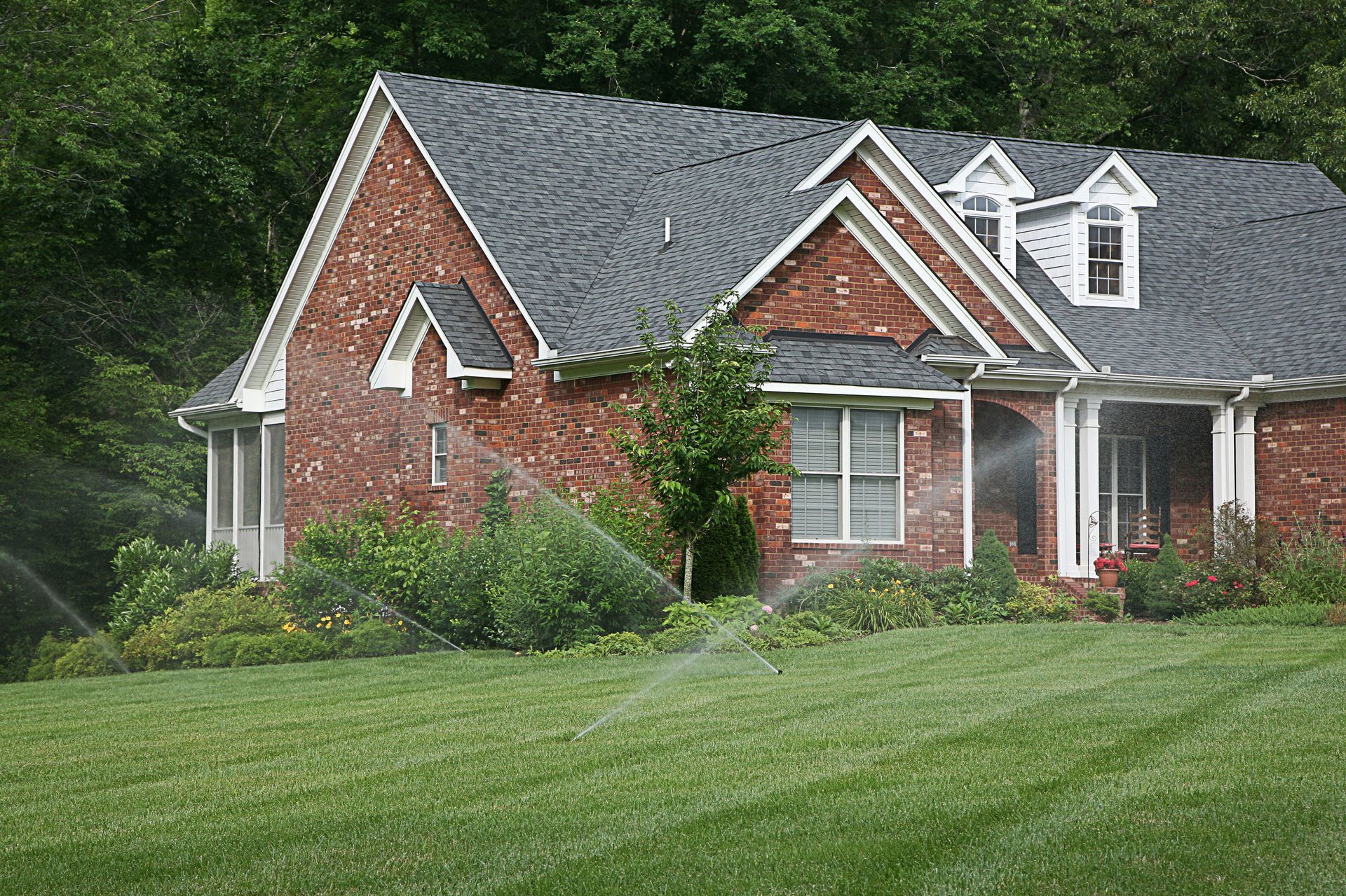 Brick house with a gray roof and white trim on a green lawn, surrounded by trees and shrubs.
