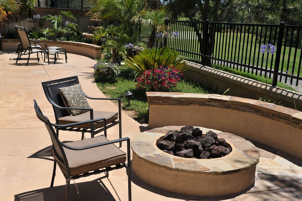 Patio seating around a circular fire pit with flowers and a black fence in the background