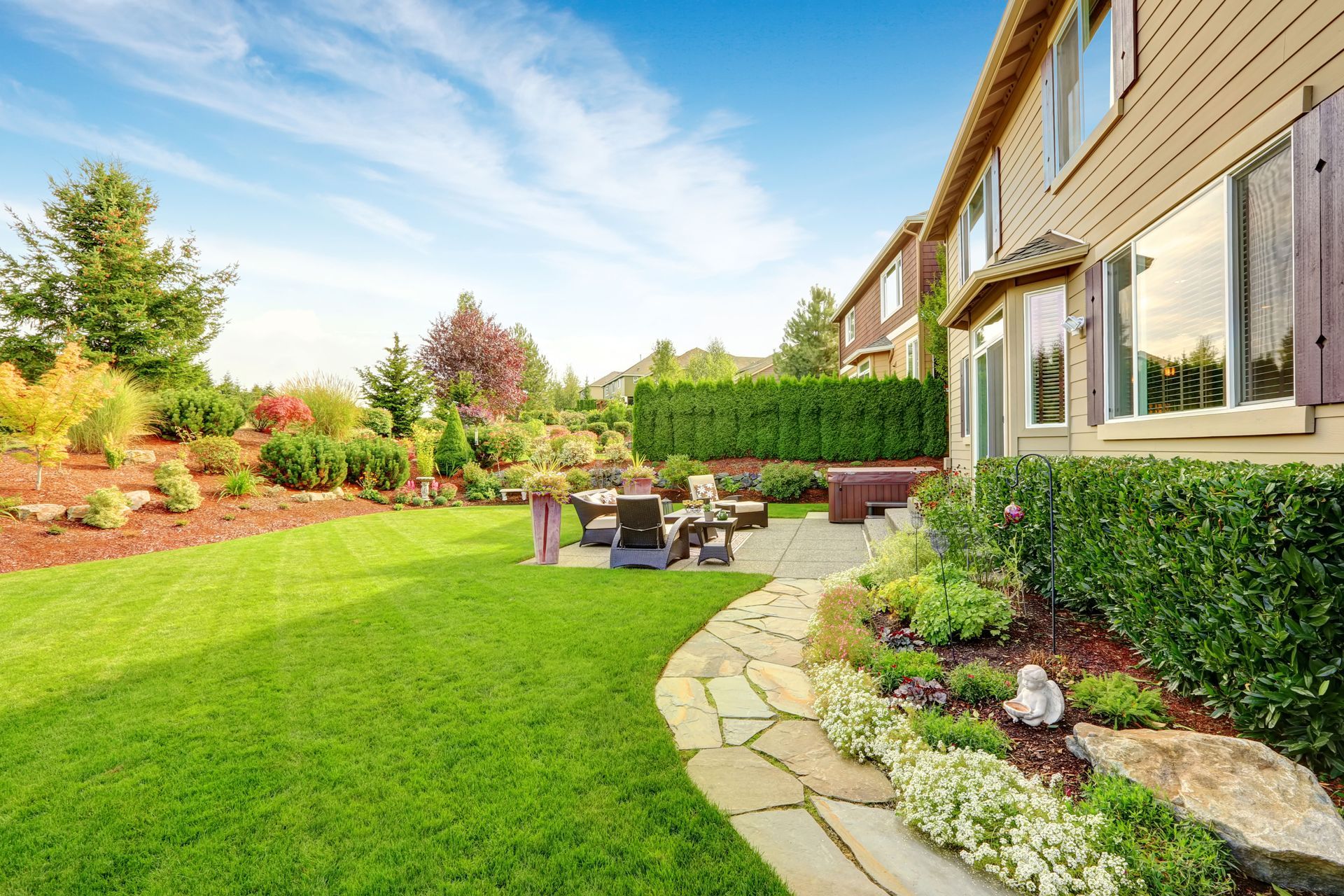 Backyard patio beside a house, with green lawn, stone path, shrubs, and outdoor seating under a blue sky
