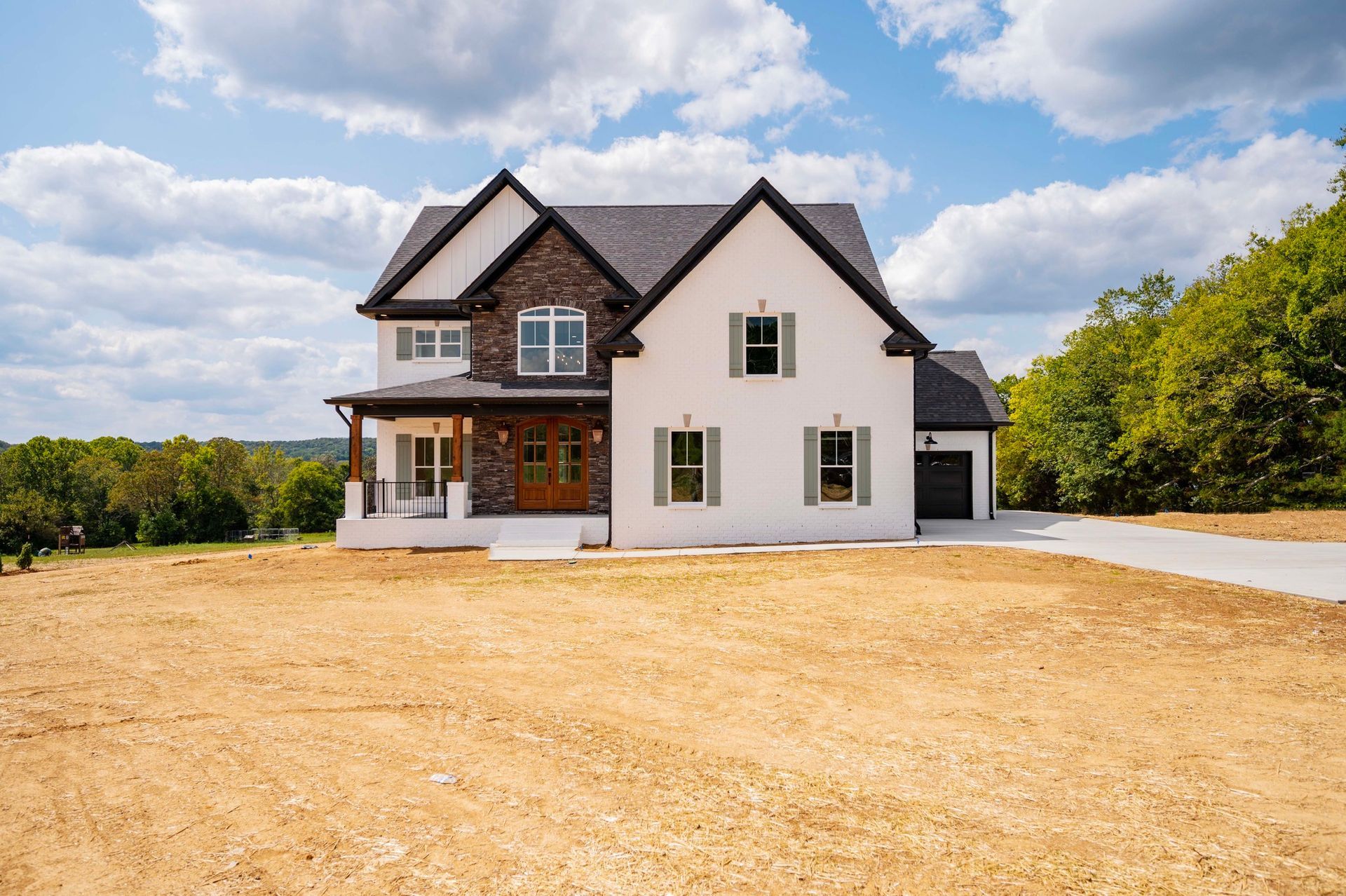 White and stone house with brown door, green shutters, and a garage, set on a brown, cleared lot under a blue sky.
